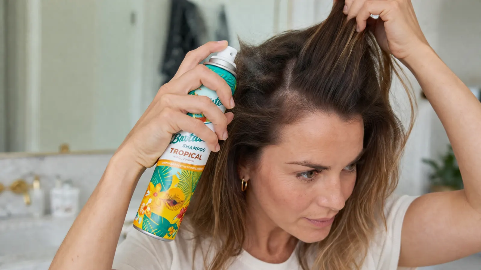 Hand holding the Batiste Tropical dry shampoo can and spraying it at the roots, sectioning hair with the other hand
